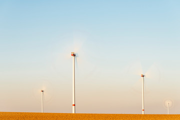 4 white wind turbines with a red stripe stand on a freshly plowed field. The rotors turn in the wind.