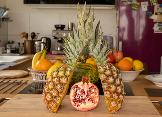 Close-up view of two halve pineapple fruit and a halve Pomegranate fruit on a cutting board in a domestic kitchen with other varieties of fruit in the background