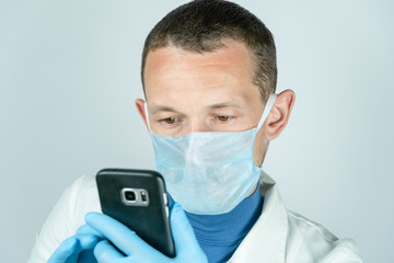 Fototapeta na wymiar Portrait of a male doctor in a medical gown, mask and gloves holding a black mobile phone and looking into it