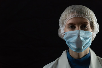 Young female doctor in medical mask, medical gown and cap on black background with copy space