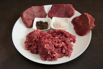 Raw fresh home-made ground beef in a white bowl, chunks of beef meat, salt and pepper close-up on a dark concrete background. Selective focus.