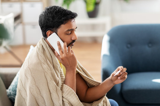 People, Health And Fever Concept - Sick Indian Man In Blanket With Thermometer Calling On Smartphone At Home