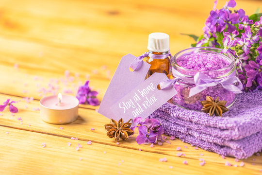 Stay at home and relax text phrase on label sticker. Spa still life with violet oil, towel and perfumed candle on natural wood table surface. Quarantine coronavirus life concept