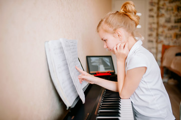 schoolgirl studying notes and playing the classic digital piano while watching an online lesson on...