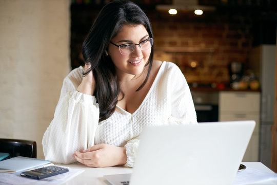 Confident attractive young plus size female in white blouse using laptop for distant work, sitting in home interior. Chubby student girl doing homework, surfing internet on portable computer - Powered by Adobe
