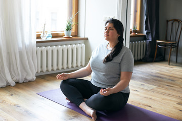 Portrait of plus size young Caucasian woman in sportswear sitting on floor using yoga mat while...