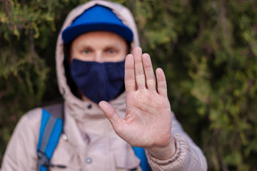 Portrait of a man wearing dark blue protective face mask against COVID-19 coronavirus disease. Stop epidemic hand gesture. Male tourist in snapback and hood on the green street during virus pandemic.