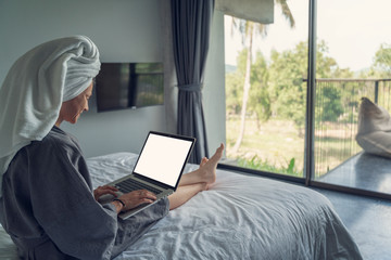 Young beautiful young woman in a white shirt sits on a white bed in a hotel room and works on a laptop