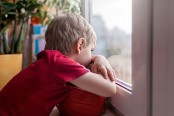 Cute sad little kid sitting on the windowsill and looking on the street, dream during lockdown