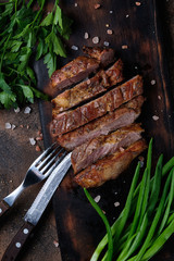 Piece of grilled meat with parsley and leek on cutting board and cutlery on dark background. Top view