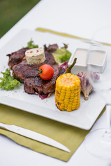Beef steak on a plate with butter on it, corn and cherry tomatoes and white sous.