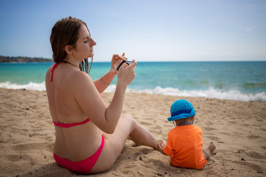 a young girl in a pink bathing suit sits on the sand of a sea beach and photographs with a smartphone her little cute baby sitting next to him in an orange T-shirt and blue hat with UV protection