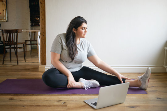 People, sports and fitness concept. Energetic plus size young woman in sneakers sitting on mat training, repeating exercises after professional instructor, watching online video lesson on laptop