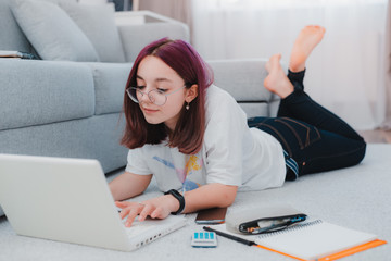 young girl teenage schoolgirl lying floor living room sofa student studying home working with her laptop doing homework.