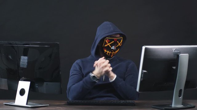 Young anonymous hacker in a mask warms his fingers on his hands to start hacking the computer.