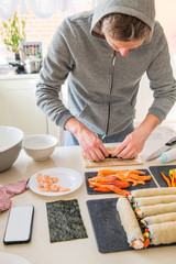Young caucasian man making sushi at home following cooking online video classes on website via smartphone. Internet technology for modern lifestyle concept. Selective focus. Verticval card. Copy space
