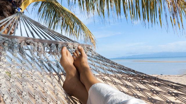 View on female feet in hammock under palm tree, swinging and relaxing  on a sand beach