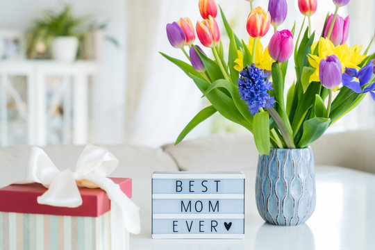 Happy Mother's Day background. Colorful spring flowers bouquet in vase, gift box with satin ribbon and lightbox with words Best mom ever on white marble table with light interior room view. Copy space