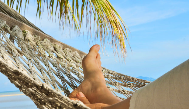 View on female feet in hammock under palm tree, swinging and relaxing  on a sand beach