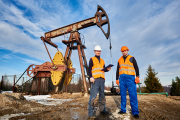 Two oil men in helmets and work vests standing on territory of oil field with oil pump...