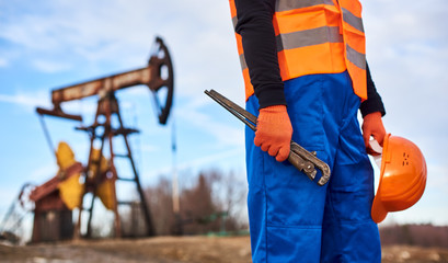Close up of petroleum operator in work orange vest holding industrial wrench and safety helmet. Oil man with instrument in hand standing on territory of oil field with pump jack on blurred background.