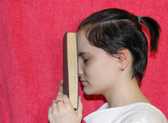 Portrait of a girl on the side, holding a thick-bound book to her forehead.