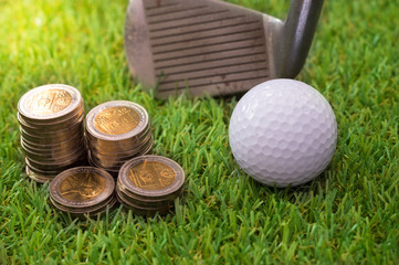 Naklejka premium Golf ball with golden coins and club on green grass.