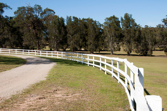 Mogo Australia, view along white wooden fence and dirt road with farm paddock in background