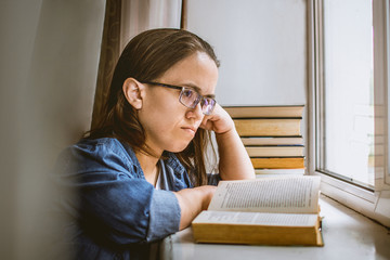 the girl with glasses looks forward and thought while reading a book near the window. a student with glasses near the window ponders the information that he read from a book