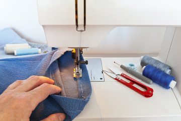 Sewing and needlework at home. Jeans hem on a sewing machine. Scissors, spools of thread and a bobbin on the desktop. Hand of seamstress in defocus