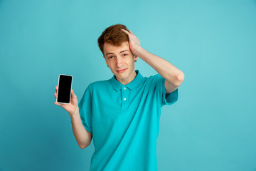 Showing blank phone screen. Caucasian young man's modern portrait isolated on blue studio background, monochrome. Beautiful male model. Concept of human emotions, facial expression, sales, ad, trendy.