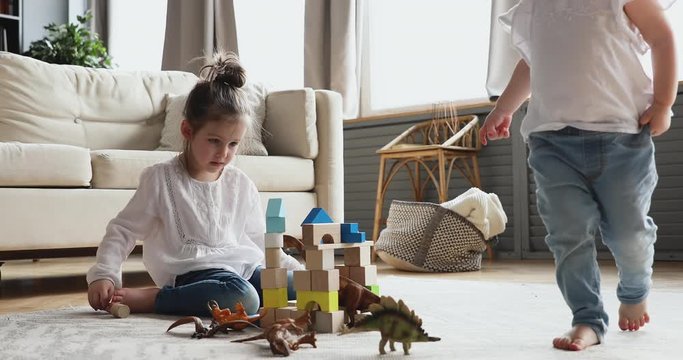 Two adorable children sisters having fun playing together helping building tower of wooden blocks sitting on warm floor carpet. Cute small preschool and toddler kids girls enjoy game in living room.