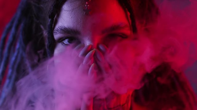 girl with dreadlocks plays with smoke through her hands in red and blue light in the house