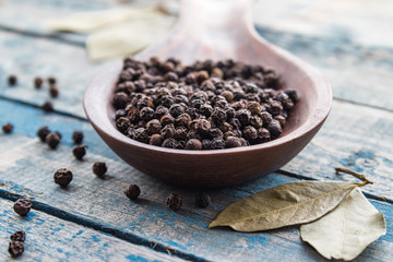 Black pepper in a large wooden spoon on a background of blue boards near a bay leaf.