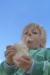 Little blond boy in green jacket biting off a piece of a bun, in the background a beautiful blue cloudless sky