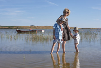 Blond mother with sunglasses, holding the sands of her son while keeping the other one in her arm, walking around in a lake with reed and boats, in Scandinavia