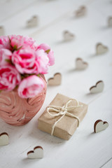 Small gift wrapped in paper with wooden hearts around and pink flowers in vase on white wooden background.