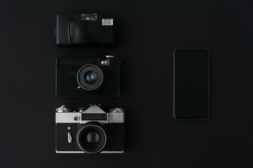 three black old film cameras lie vertically in a row on a black background in comparison with modern smartphone, space for text, history of photography and modern technologies