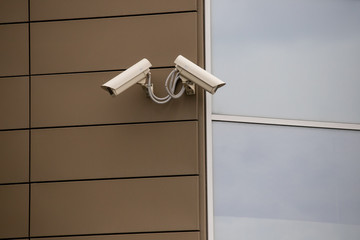 Big white surveillance camera on light beige stone column against background of public building with blue window; close-up