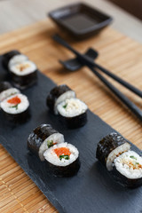 Maki sushi on a black board. Soy sauce in a plate and black chopsticks on a bamboo mat.