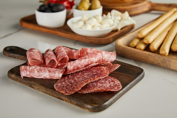 Selective focus of salami slices on board near antipasto ingredients on white