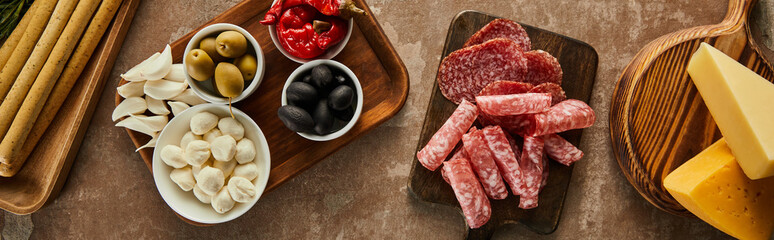Top view of cheese, salami slices and antipasto ingredients on boards on brown background, panoramic shot