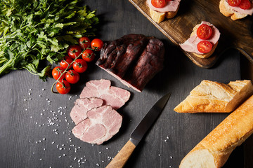 Obraz na płótnie Canvas top view of tasty ham sliced ham, cherry tomatoes, parsley, salt, knife and baguette on wooden grey table with canape