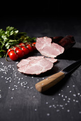 selective focus of tasty ham sliced ham, cherry tomatoes, parsley, salt, knife on wooden grey table isolated on black