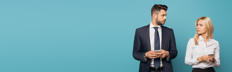 panoramic orientation of young businesswoman and businessman looking at each other while holding smartphones on blue background