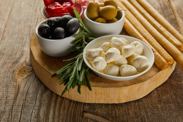 Round board with rosemary, breadsticks and bowls with antipasto ingredients on wooden background