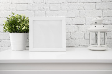White frame mockup with green plant and lantern on a white table against white brick wall.Empty poster frame mockup for design.