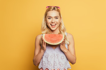 attractive blonde girl holding slice of watermelon and smiling at camera on yellow background