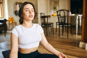 Fototapeta na wymiar Beautiful young Caucasian woman with muscular curvy body sitting in lotus posture at home keeping eyes closed, meditating during yoga practive, doing body scanning, concentrating on breathing