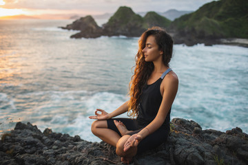 Fototapeta na wymiar Young woman practicing yoga in lotus pose at sunset with beautiful ocean and mountain view. Sensitivity to nature. Self-analysis and soul-searching. Spiritual and emotional concept.